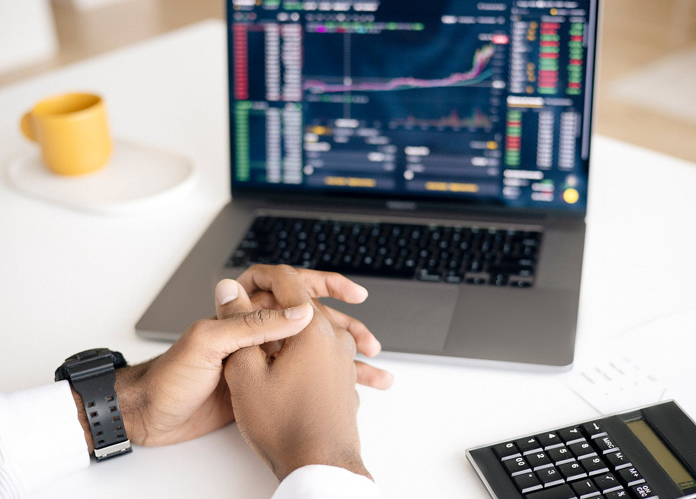 Cmputer with multicolor graphics on the screen, two man hands intertwined in each other and a calculator on the right, all on a white table, plus a yello mug on the left side of the photo, unfocused