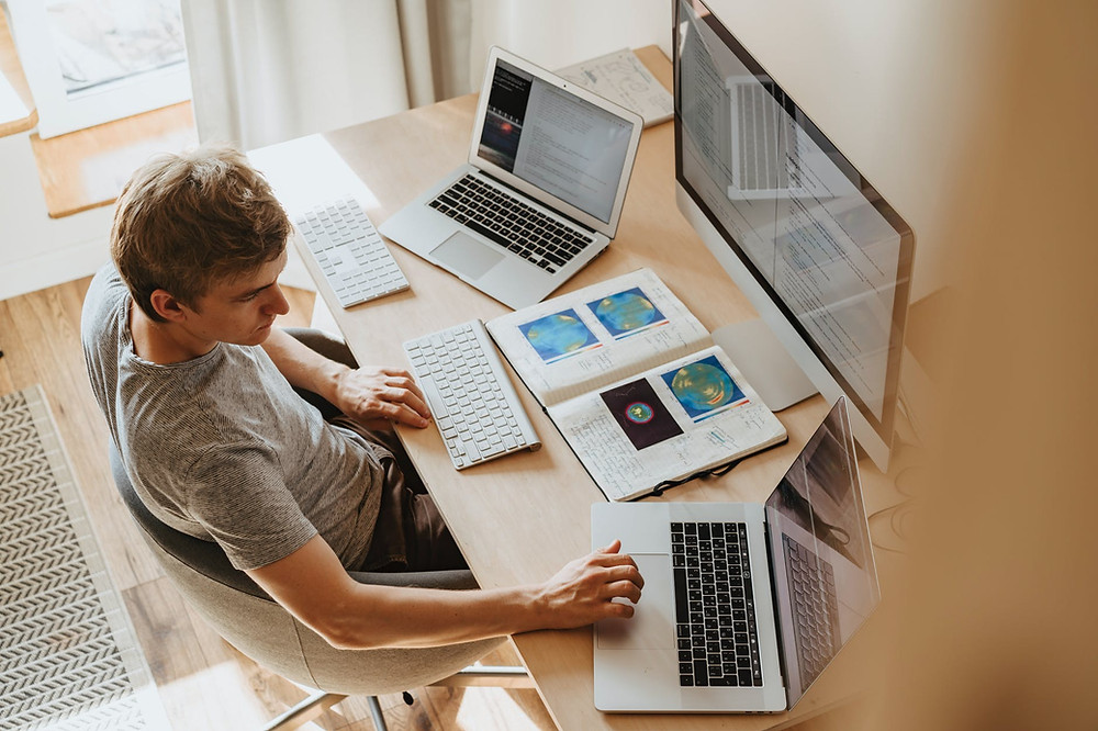 Boy sitting in front a big computer screen, with two white keyboard and a second laptop on his right and another on his left, upon a wooden table with a notebook with pictures, on it, open
