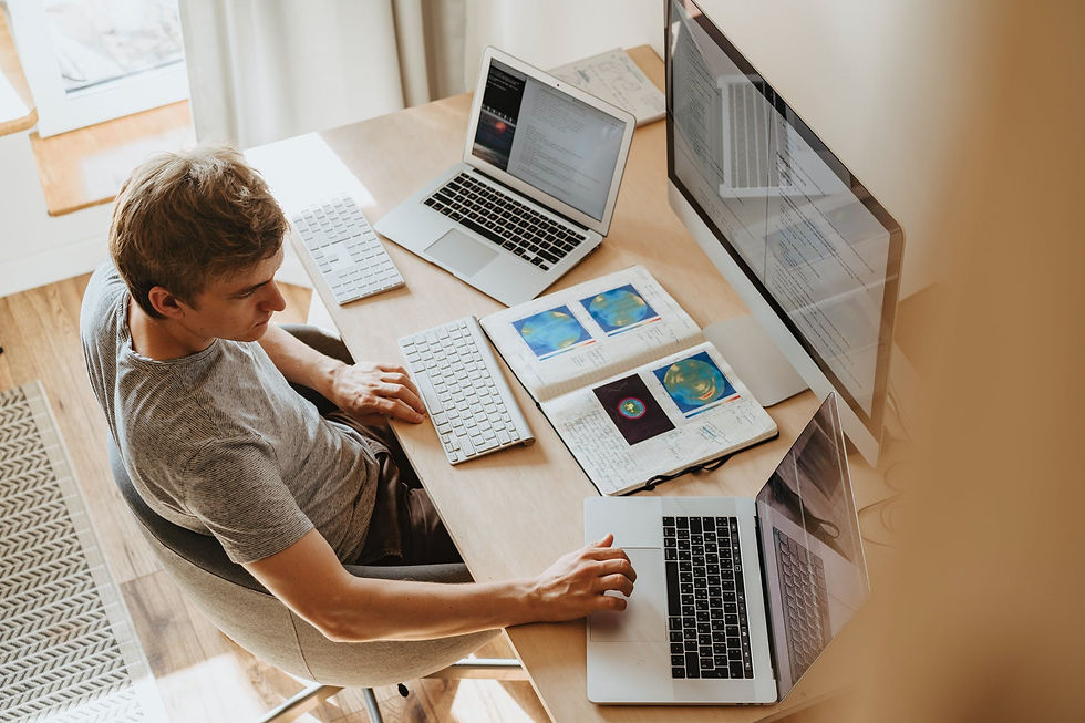 Boy sitting in front a big computer screen, with two white keyboard and a second laptop on his right and another on his left, upon a wooden table with a notebook with pictures, on it, open