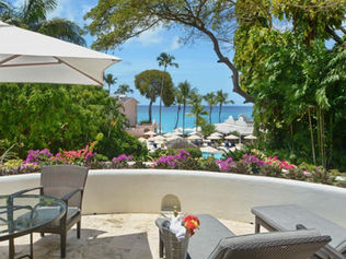 Terrasse de jardin avec table en verre, chaises et parasol. Vue sur une piscine entourée de palmiers et la mer turquoise en arrière-plan.