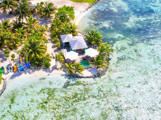 île tropicale avec maisons en bois, piscine, parasols blancs et palmiers. Kayaks colorés alignés sur le sable près de l'eau turquoise.
