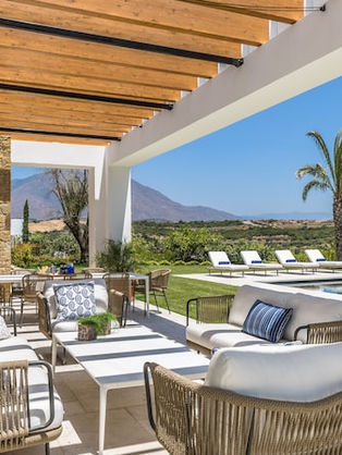 Terrasse ensoleillée avec canapés beiges, piscine bleue, palmiers, chaises longues blanches, montagnes en arrière-plan. Ambiance détendue.