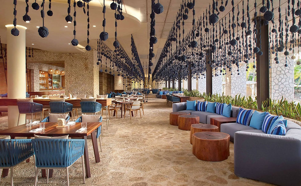 Restaurant élégant avec tables en bois, chaises bleues, canapés gris et coussins à rayures. Décorations suspendues, ambiance chaleureuse.