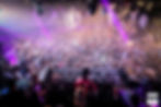 DJ dirige une foule déchaînée dans une boîte de nuit. Lumières violettes et confettis volants. Ambiance festive et électrisante.