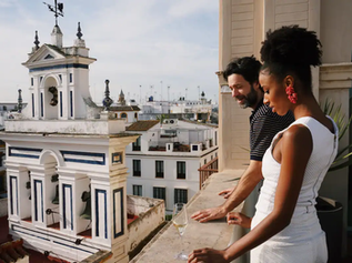 Un homme et une femme sur un balcon, regardant une ville avec une tour blanche. La femme porte une robe blanche. Ambiance détendue.