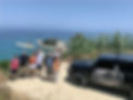 a goup of eight people are possing in front of a sea front landscape with limestone cliffs and mediteranean bamboo plants, a black jeep with the logo "x adventure" is parked on their left hand side
