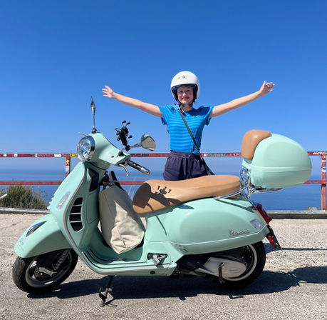 Young smilling lade possing with wide open hands right behing a Vespa scooter
