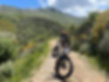 2 bicycles in front of one of them is a rider possing for the photo wearing a red helmet, is on a gravel road, behind him is the mount pantokrator, the area have many bushes no tall trees, there are yellow flowered bushes in many spots blending with the vivid green coloured grass and the sage plants