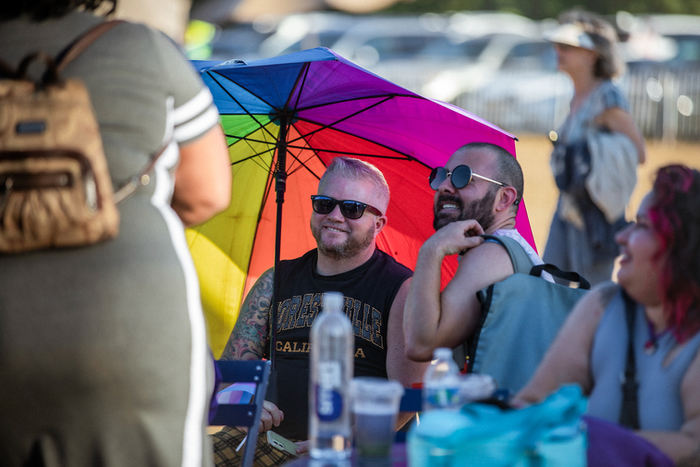 group of two masc people and leaders of pi wear sunglasses and smile at a group of people outside of the frame. they are sitting under an umbrella of vibrant rainbow colors.