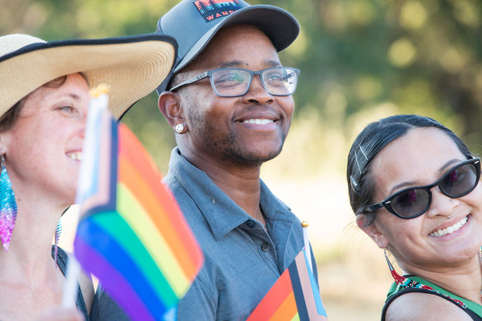 group of three people smile and hold inclusive pride flags on a sunny day