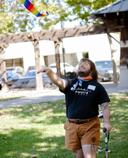 Joy wears a black shirt with a PI rainbow logo and stands on green grass with their arm raised and poised with a rainbow kite on a sunny day at at a social saturday event windsor town green.