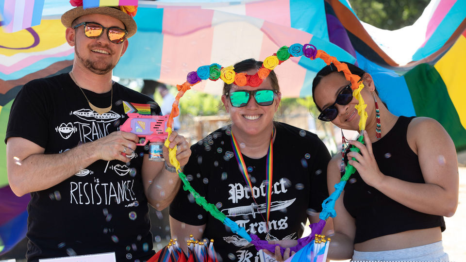 drew, jess and marian are smiling and wearing sunglasses at a pride event while holding a rainbow-colored heart shape that frames jess' face. they stand under a billowing pride flag.