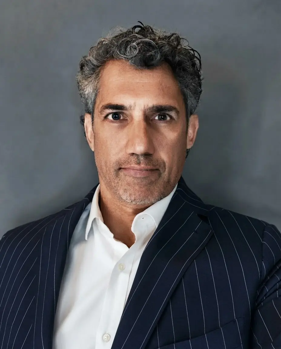 Studio portrait of a middle-aged man with curly salt-and-pepper hair, wearing a pinstripe blazer and white shirt. The mood is professional and poised against a neutral grey backdrop.