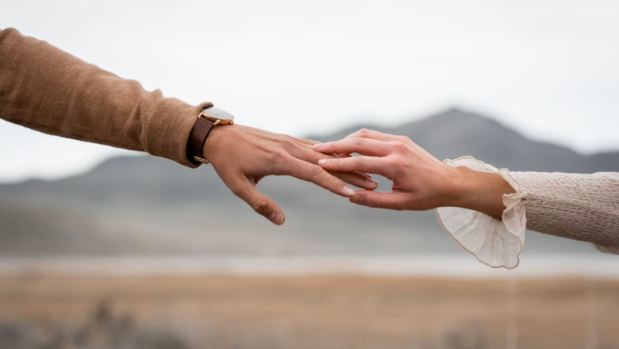 Two hands touch fingertips outdoors, with blurred mountains in the background and a soft, romantic mood.