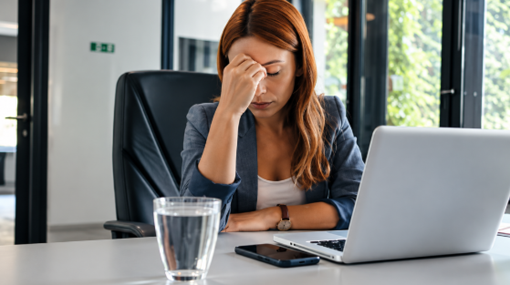 Stressed woman in an office rests her head on her hand beside a laptop, phone, and glass of water, looking tired.