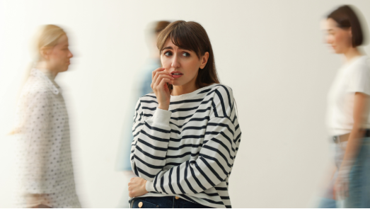 Anxious woman in striped shirt stands in foreground, biting her nail as blurred people walk past against a plain wall.
