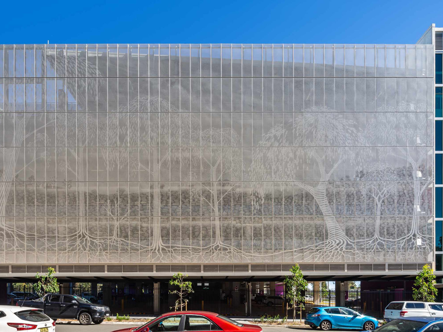 Edmondson Park Commuter Car Park - Modern building with a translucent tree mural facade, blue windows, parked cars below, and clear blue sky.