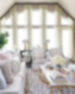 Elegant living room with light beige furniture, pastel pillows, and gold-accent coffee table. Large windows with sheer curtains, airy and calm.