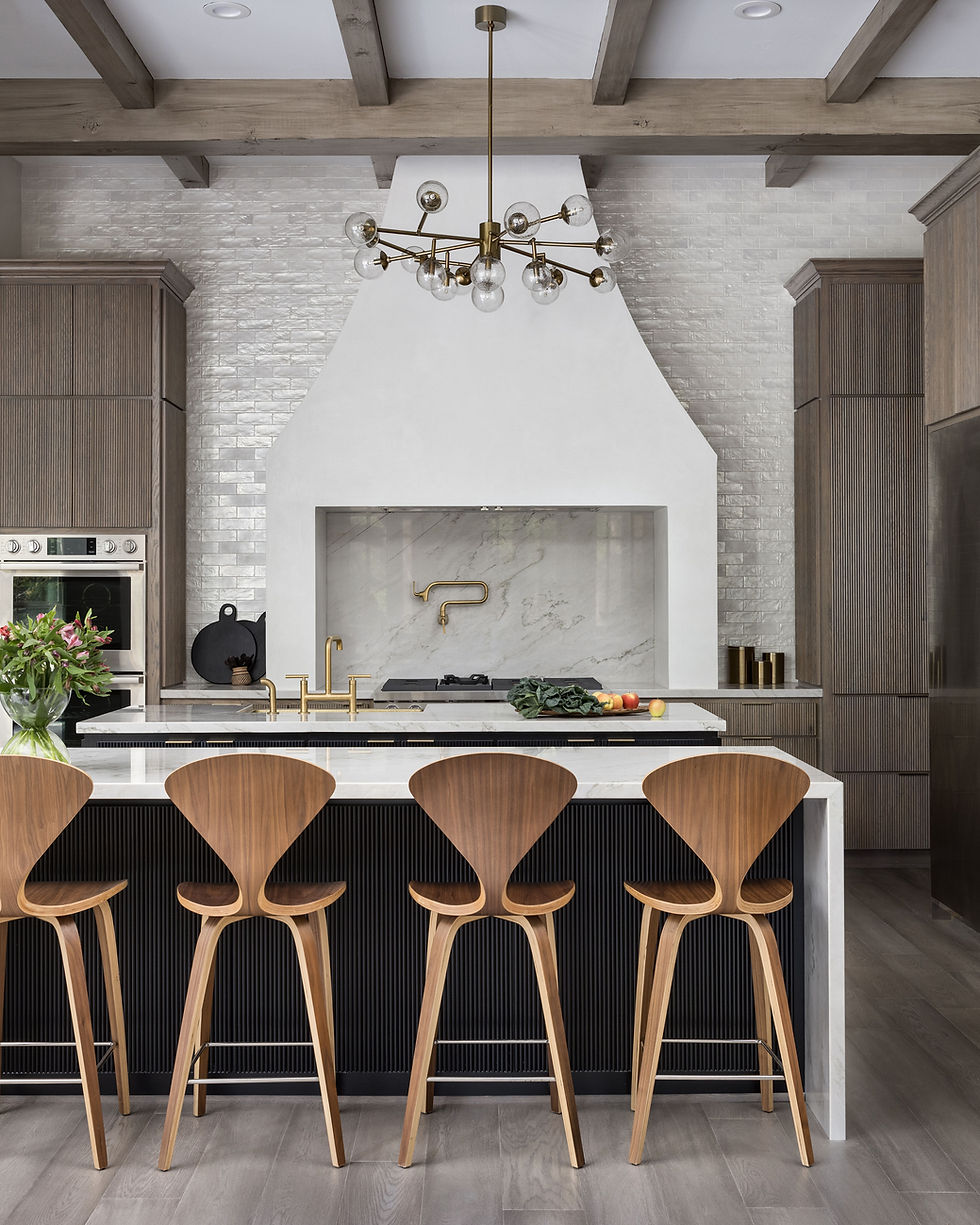 Modern kitchen with wooden bar stools at a marble island, brass fixtures, flowers, and fresh produce. Chic chandelier and wooden beams above.