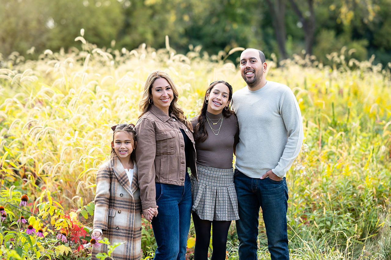 A family of 4 in neutral brown clothing poses for a family portrait in a field of wildflowers during fall portraits at the River View Farmstead in Naperville, IL