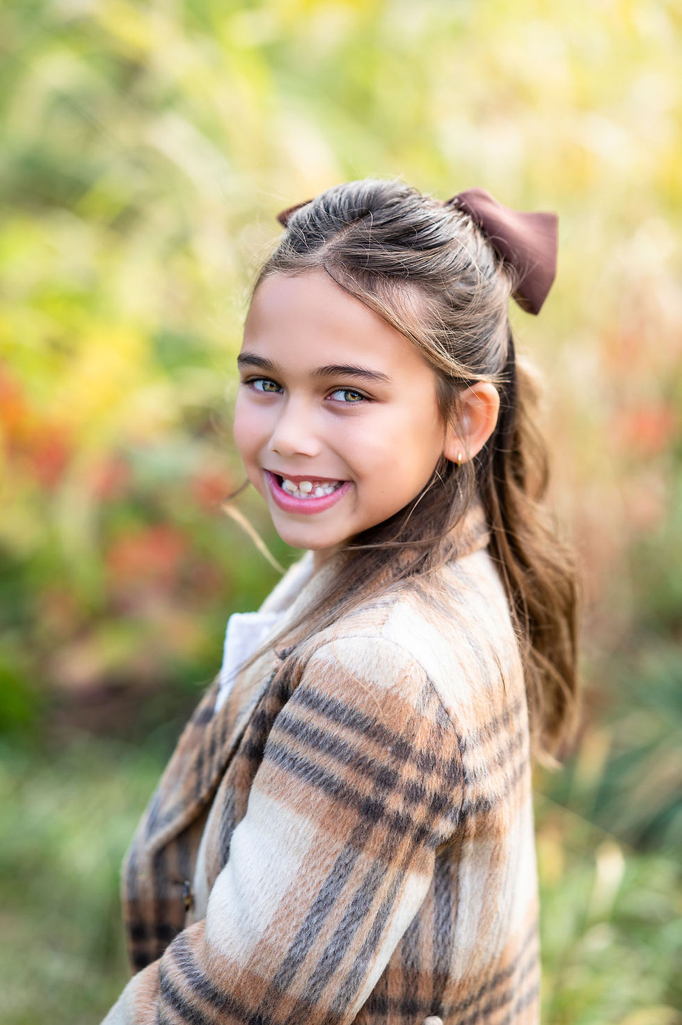 Smiling young girl outdoors in a plaid coat with a brown hair bow, against a soft green-and-gold blurred background at the River View Farmstead in Naperville, IL