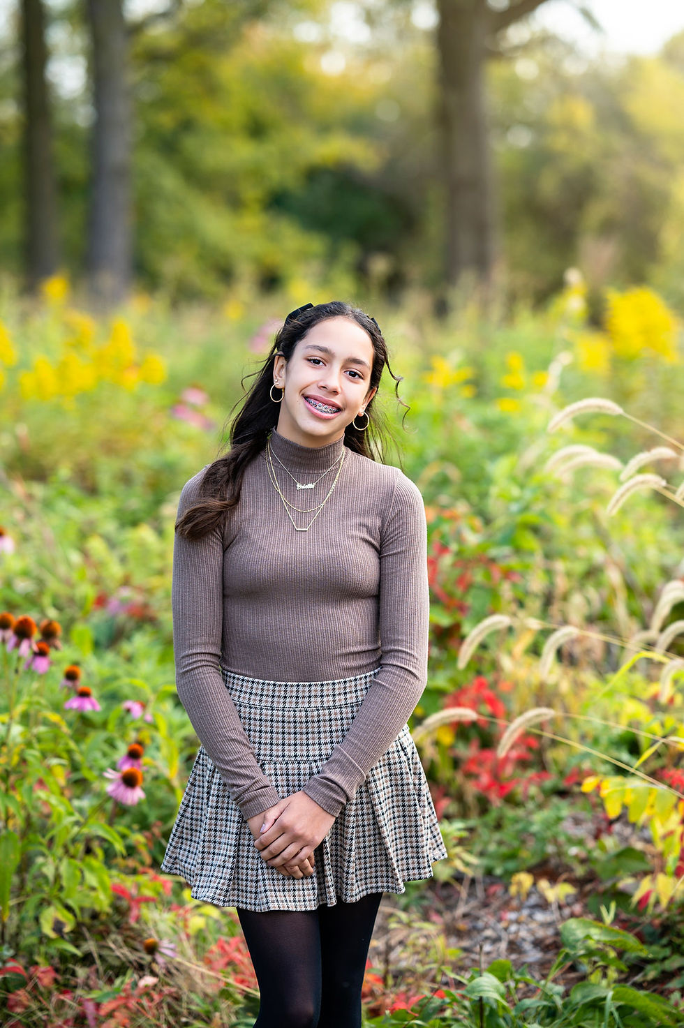 A teenage girl in a brown sweater and grey plaid skirt poses in front of the wildflowers on the riverbank of the DuPage River at the River View Farmstead in Naperville IL