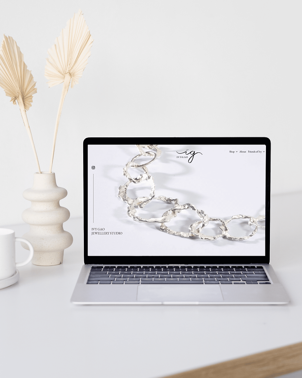 Minimal desk with laptop showing Ivy Gao Jewellery Studio website, silver chain jewelry, and a cream vase with dried leaves.