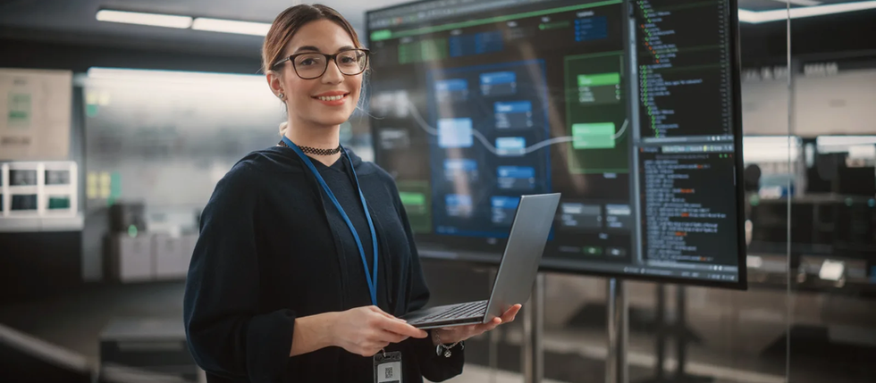 A person with glasses, smiling and holding a laptop, stands in front of a large screen displaying code in a modern, dimly lit office.