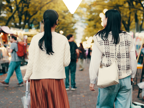 Two women walk away through a tree-lined outdoor market at dusk, with warm lights, stalls, and autumn leaves around them.