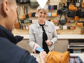 Smiling shop clerk hands a blue card reader to a customer in a purse shop, with colorful bags on the wall and Driftscape logo.