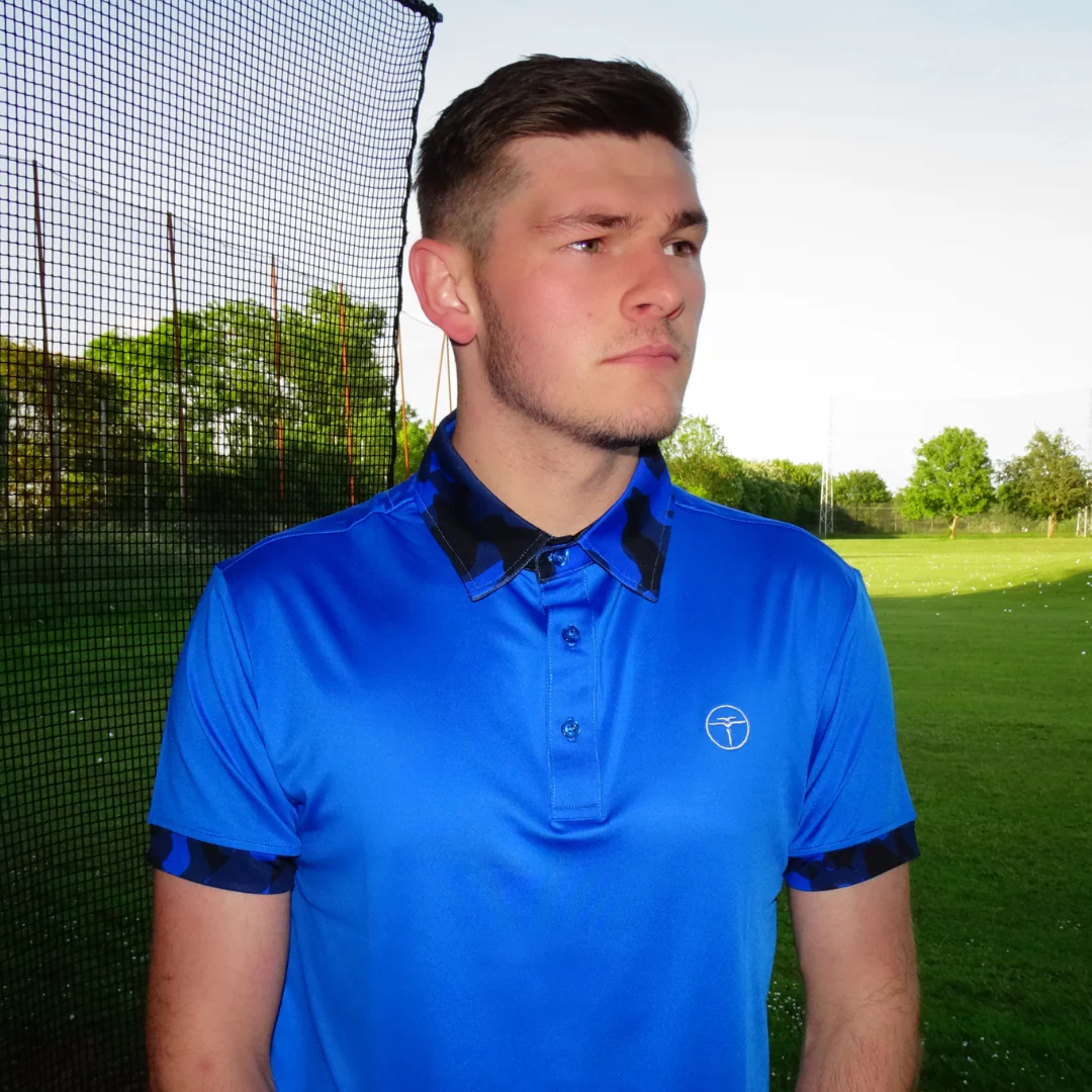 A young man wearing a bright blue polo shirt stands outdoors on a grassy field near a net, with trees and blue sky in the background.