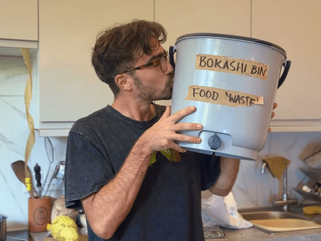 Man in a kitchen kisses a gray bin labeled "Bokashi Bin Food Waste," showing affection. Kitchen utensils and light cabinets in background.