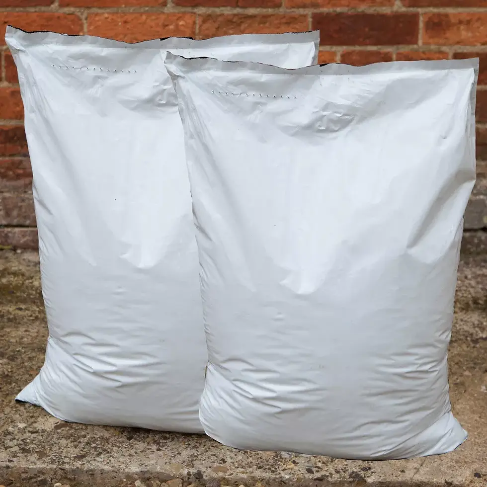 Two large white sacks of commercial bagged compost stand on a stone surface against a brick wall. Simple and utilitarian setting.