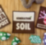 Generation Soil x Rebel Patch sew on patches on wooden table surrounded by handmade mushrooms and crochet worm