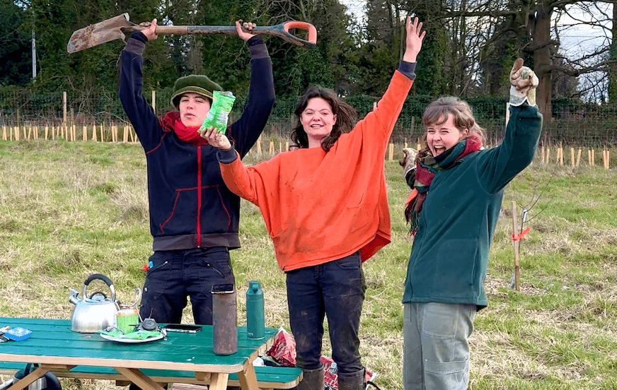 Three people celebrate outdoors; one holds a shovel, another has a packet. They smile energetically in a grassy field with trees behind.