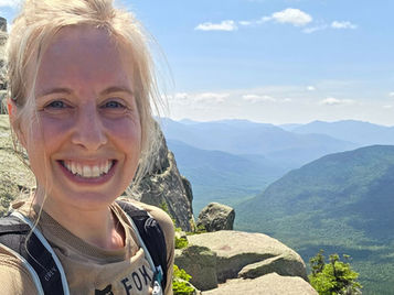 Smiling Amanda McKeen takes a selfie on a rocky mountain ledge, with blue skies, layered peaks, and FOX on her tan shirt.
