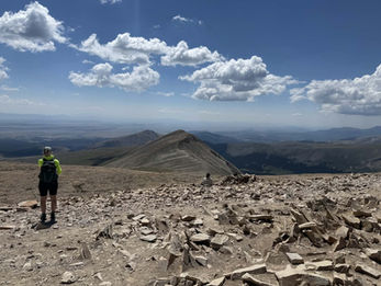 Hiker in neon jacket stands on a rocky mountain summit while another sits nearby, overlooking vast peaks under a bright blue cloudy sky.