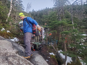 Hikers with backpacks climb a rocky, snowy forest trail; a man in a blue jacket and neon hat leads under tall evergreens.