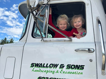 Two smiling children in a truck's cab, with clear blue sky above. The truck has "Swallow & Sons Landscaping & Excavation" text.