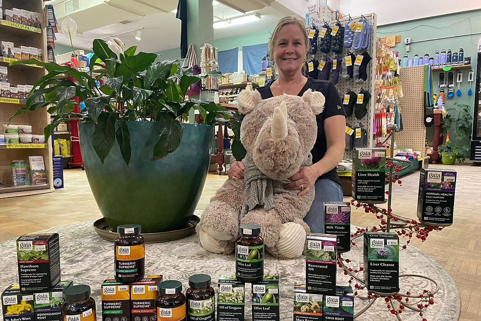 Barbara Emmons sits with a plush rhino in The Healthy Rhino, surrounded by plant supplements on display. A large green plant is beside her.