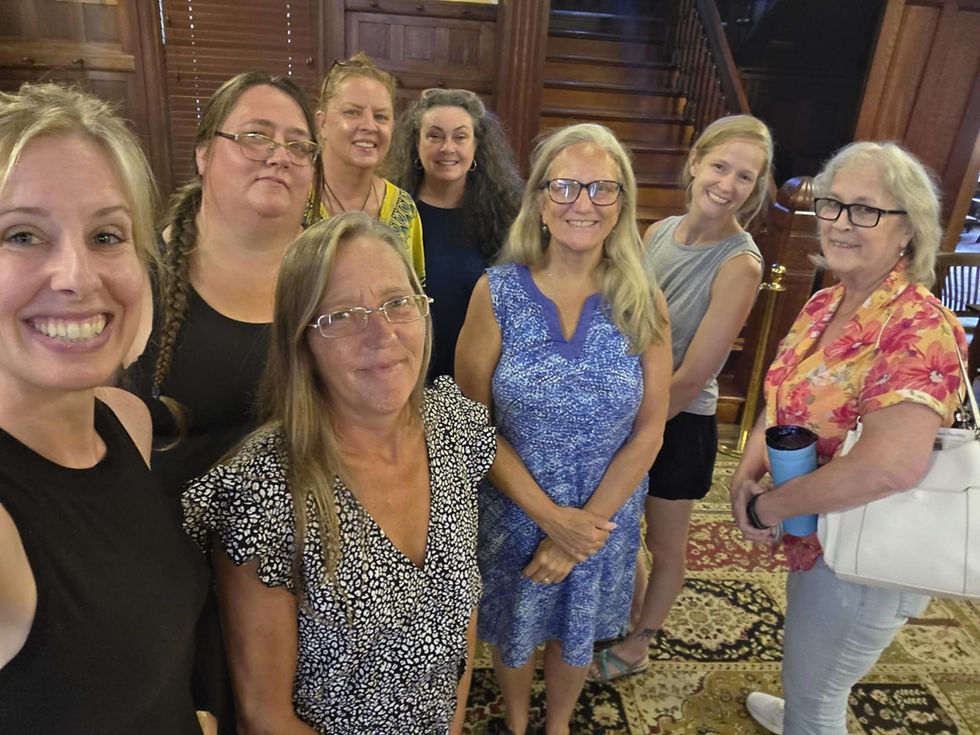 Smiling group selfie of seven women in a wood-paneled hall with stairs and patterned carpet; casual dresses, one holding a blue tumbler