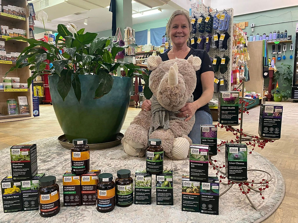 Barbara Emmons sits with a plush rhino in The Healthy Rhino, surrounded by plant supplements on display. A large green plant is beside her.