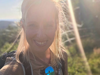 Amanda McKeen on a sunny trail, backpack on, sunglasses hanging from shirt, with bright lens flare and green hills.