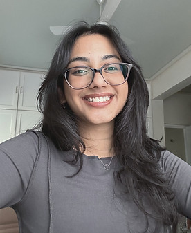 Photo of Siya. She wear a grey blouse, with dark-coloured spectacles, a small silver necklace, and tiny drop-earrings. Her hair is left open and draped over her left shoulder, and she is smiling into the camera with her teeth showing