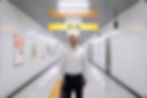 The character 'the smiling man' from new horror film 'Exit 8' stands in the centre of the image, smiling uncannily down the lens.