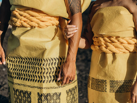 Two people in traditional Hawaiian yellow patterned garments pose confidently outdoors against a blue sky, with volcanic rocks in the background.