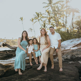 Family of five smiling on a beach, sitting on driftwood. They're dressed in pastel colors. Palm trees and sunlight in the background. Hawaii beach photographer