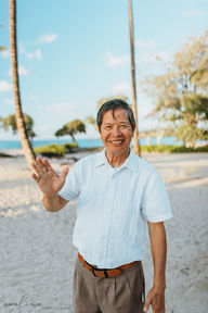 Family of ten smiling on a beach, wearing light, patterned outfits. Rocky shoreline and ocean in the background. Warm, joyful mood. Hawaii sunset beach photo session
