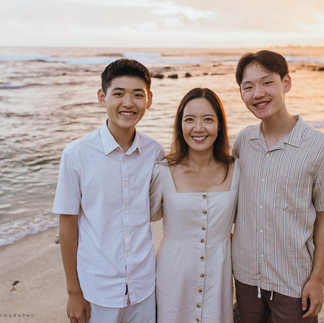 A family of six stands on a beach at sunset, smiling. The sky is orange, and the ocean is calm. They're dressed in light, casual attire.