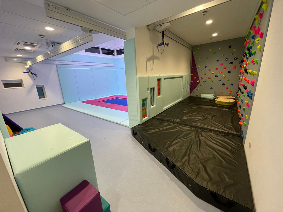 A large theatre lit trampoline area with hoist system above and to the right a bespoke climbing wall with padded floor and tactile wall panels in pale blue vinyl with the padded data cabinet in the foreground
